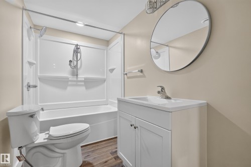 Bathroom featuring a white vanity with integrated sink, chrome fixtures, a round mirror, and wood-finish flooring - 7014 Chivers Loop Sw, Edmonton, AB 