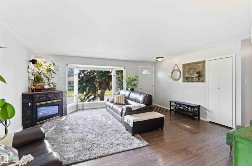Bright living area featuring wood-finish flooring, a bay window, a built-in electric fireplace with stone-look surround, a front entry door with an arched window, and a white bi-fold closet door - 5208 105 Street Nw, Edmonton, AB - Indoor Photo Showing Living Room With Fireplace