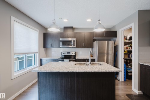 Modern kitchen featuring a central island with dual pendant lighting, stainless steel appliances, dark wood-finish cabinetry, light speckled countertops, and a full pantry - #28 2215 24 St Nw, Edmonton, AB - Indoor Photo Showing Kitchen With Double Sink With Upgraded Kitchen