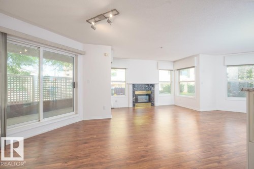 Spacious living area featuring wood-finish flooring, a fireplace with a dark stone surround, and multiple windows - #108 12028 103 Av Nw, Edmonton, AB 