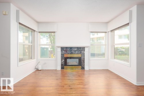 Wood-finish flooring, central fireplace with dark stone surround, white mantelpiece, and multiple windows - #108 12028 103 Av Nw, Edmonton, AB 