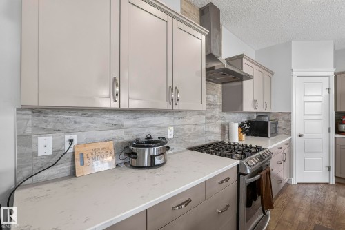 Contemporary kitchen featuring light-toned cabinetry, a stainless steel gas range, a coordinating stainless steel range hood, stone-finish countertops, and a full-height tile backsplash - 923 174St Sw, Edmonton, AB 