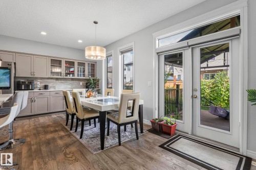 Kitchen and dining area featuring wood-finish flooring, light gray cabinetry with glass-front uppers, an integrated breakfast bar, and a contemporary chandelier - 923 174St Sw, Edmonton, AB 