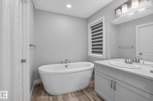 Freestanding oval bathtub with chrome fixtures, wood-finish flooring, white vanity with integrated sink, rectangular mirror, and window with horizontal blinds - 923 174St Sw, Edmonton, AB 