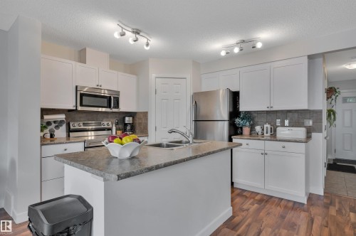 Kitchen featuring white cabinetry, stainless steel appliances, a center island with a sink, wood-finish flooring, and track lighting - 17321 8A Avenue Sw, Edmonton, AB - Indoor Photo Showing Kitchen With Double Sink