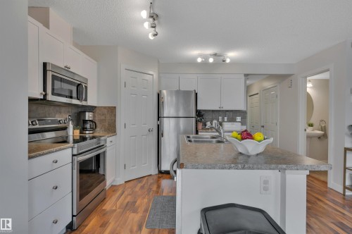 Kitchen featuring stainless steel appliances, wood-finish flooring, white cabinetry, a stone-finish countertop island, and tile backsplash - 17321 8A Avenue Sw, Edmonton, AB - Indoor Photo Showing Kitchen With Stainless Steel Kitchen With Double Sink