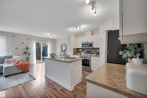Kitchen island with an undermount sink and stone-look countertop, white cabinetry with stainless steel appliances, and wood-finish flooring - 17321 8A Avenue Sw, Edmonton, AB - Indoor Photo Showing Other Room