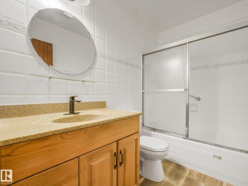 Bathroom featuring a wood-finish vanity with an integrated sink, a granite-look countertop, a modern single-handle faucet, a shower/tub combination with sliding glass doors, and tiled walls - 16428 88 Avenue Nw, Edmonton, AB - Indoor Photo Showing Bathroom