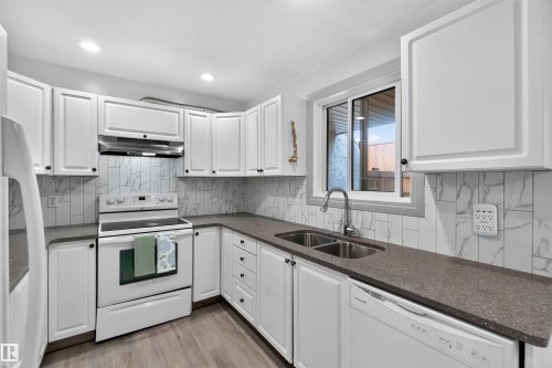 White cabinetry with dark hardware, stone-finish countertops, stainless steel double basin sink, white electric range, and wood-finish flooring - #28 3645 145 Av Nw, Edmonton, AB - Indoor Photo Showing Kitchen With Double Sink