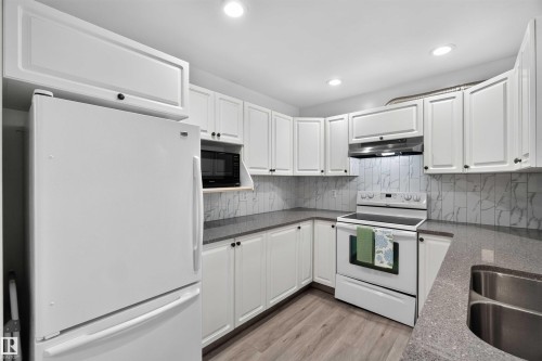 Kitchen featuring white cabinetry with dark hardware, a stainless steel double sink, light-toned wood-finish flooring, and recessed lighting - #28 3645 145 Av Nw, Edmonton, AB - Indoor Photo Showing Kitchen With Double Sink