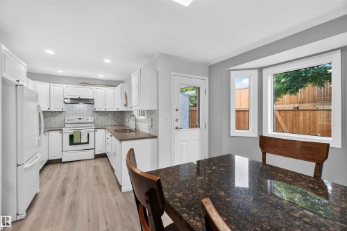 Kitchen featuring white cabinetry, dark countertops, a white appliance suite, wood-finish flooring, and a bay window - #28 3645 145 Av Nw, Edmonton, AB - Indoor Photo Showing Kitchen With Double Sink