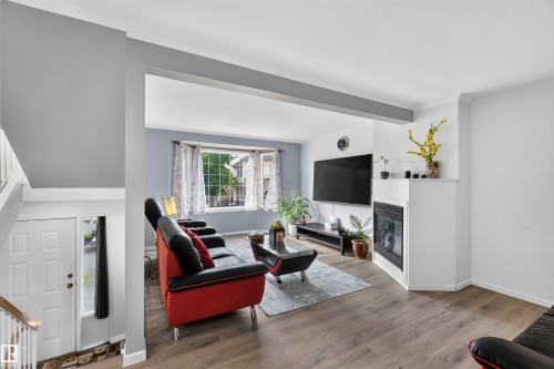 Wood-finish flooring, a white fireplace with a dark insert, a prominent bay window, and a white front door with a side light - #28 3645 145 Av Nw, Edmonton, AB - Indoor Photo Showing Living Room With Fireplace