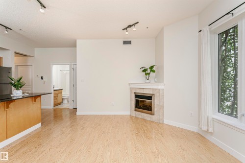 Light wood-finish flooring throughout, corner fireplace with tile surround, white window trim, and track lighting - #219 10333 112 St Nw, Edmonton, AB - Indoor With Fireplace