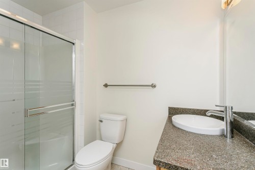 Glass-enclosed shower with white tiled surround, toilet, vanity with integrated basin, polished chrome faucet, and mirrored wall - #219 10333 112 St Nw, Edmonton, AB - Indoor Photo Showing Bathroom