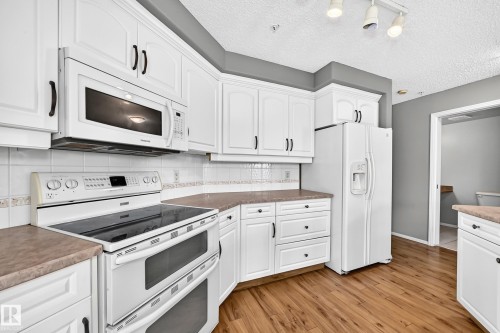 White cabinetry with dark hardware, stone-finish countertops, tile backsplash, built-in microwave, and wood-finish flooring - #331 9704 174 St Nw, Edmonton, AB 