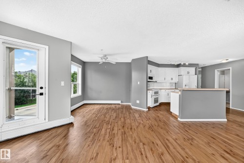 Open-concept living area featuring wood-finish flooring, a ceiling fan, and abundant natural light from the large windows and glass-paneled door - #331 9704 174 St Nw, Edmonton, AB 