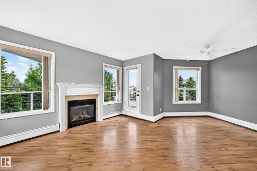 Living area featuring wood-finish flooring, a white mantel fireplace, multiple windows, a white ceiling fan, and a glass-paneled exterior door - #331 9704 174 St Nw, Edmonton, AB 