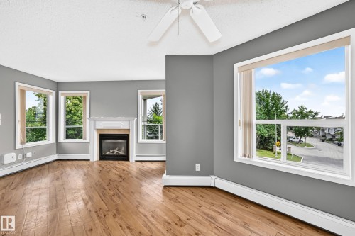 Wood-finish flooring, white trim, multiple windows with blinds, a built-in fireplace with a white mantel, and a ceiling fan - #331 9704 174 St Nw, Edmonton, AB 