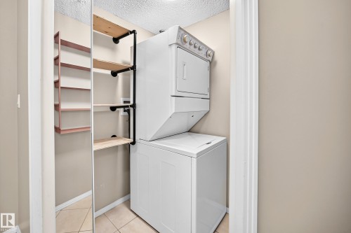 Stacked washer and dryer unit, tiled flooring, open shelving with wood shelves and black pipe supports, additional built-in shelving, and a mirrored closet door - #331 9704 174 St Nw, Edmonton, AB 