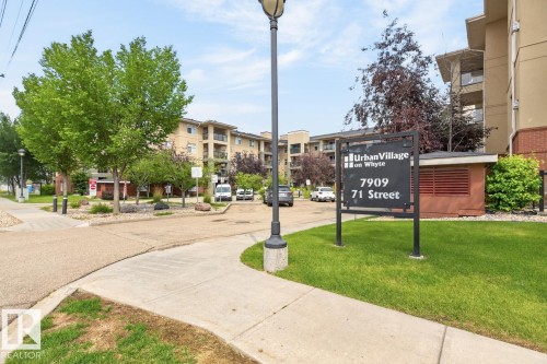 Building exterior featuring a neutral tone facade with balconies, surrounded by mature trees and landscaped grounds - #210 7909 71 St Nw, Edmonton, AB - Outdoor
