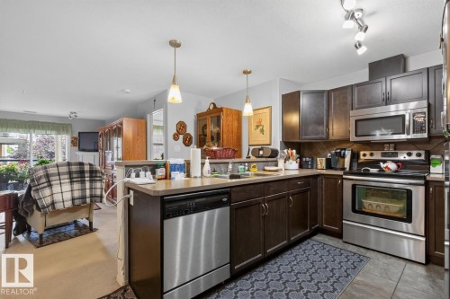 Kitchen featuring dark wood-finish cabinetry, stainless steel appliances, pendant lighting, and a neutral countertop - #210 7909 71 St Nw, Edmonton, AB - Indoor Photo Showing Kitchen With Double Sink
