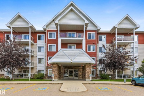 Multi-story building featuring red and beige siding, gabled rooflines, multiple private balconies with glass railings, an inviting stone-clad entrance, and paved parking - #117 2203 44 Av Nw, Edmonton, AB - Outdoor With Facade