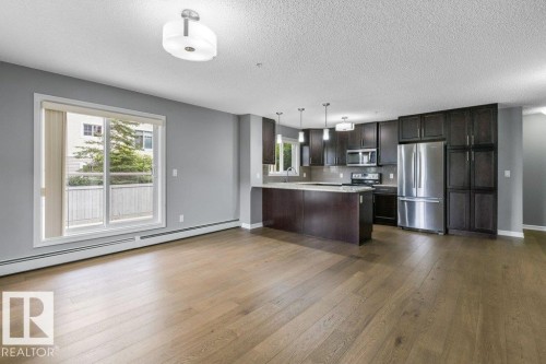 Open-concept living space with wood-finish flooring, a sliding glass door, and an integrated kitchen featuring dark wood cabinetry, stainless steel appliances, and pendant lighting - #117 2203 44 Av Nw, Edmonton, AB - Indoor Photo Showing Kitchen