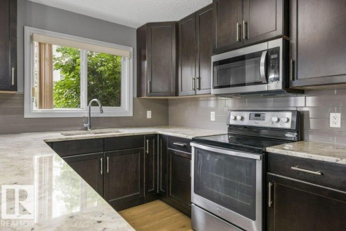 Dark wood-finish cabinetry, light-toned countertops, stainless steel appliances, and a tile backsplash - #117 2203 44 Av Nw, Edmonton, AB - Indoor Photo Showing Kitchen With Upgraded Kitchen
