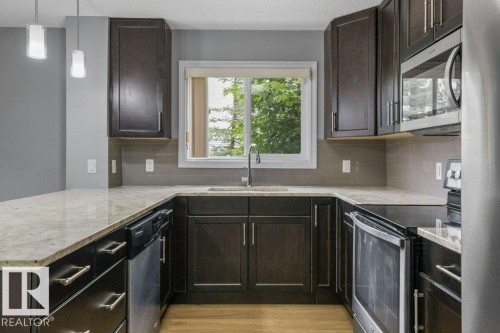 Modern kitchen featuring dark cabinetry with brushed metal hardware, light stone countertops, and stainless steel appliances including a built-in microwave - #117 2203 44 Av Nw, Edmonton, AB - Indoor Photo Showing Kitchen With Upgraded Kitchen