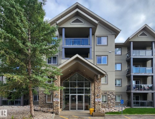 Modern multi-unit residential building featuring a striking stone and stucco exterior with large glass entrance doors and transom windows - #128 279 Suder Greens Dr Nw, Edmonton, AB - Outdoor With Balcony With Facade