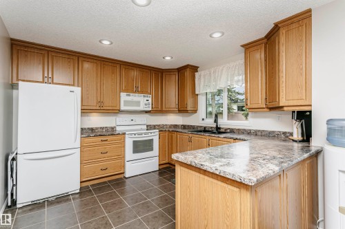 Kitchen featuring wood-finish cabinetry, integrated white appliances, L-shaped countertop, recessed ceiling lighting, and dark-toned floor tiles - #42 9630 176 St Nw, Edmonton, AB - Indoor Photo Showing Kitchen