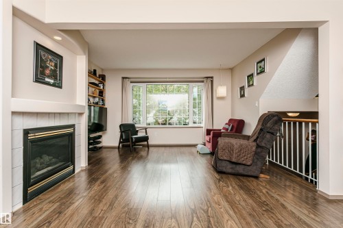 Fireplace with white tile surround, wood-finish flooring, large multi-pane window, built-in shelving, and recessed lighting - #42 9630 176 St Nw, Edmonton, AB - Indoor Photo Showing Living Room With Fireplace