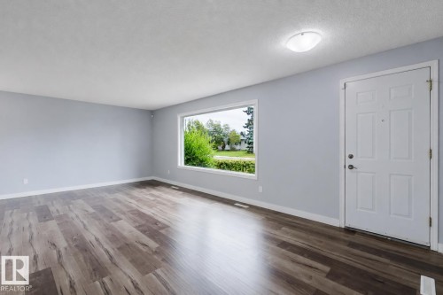 Expansive room featuring wood-finish flooring, a large window, a white entry door, and a flush-mount ceiling light - 11420 46 Avenue Nw, Edmonton, AB - Indoor