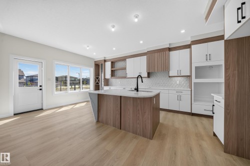 Modern kitchen featuring a curved island with integrated sink, white and wood-finish cabinetry, and a geometric tile backsplash - 8915 Mayday Way Sw, Edmonton, AB - Indoor Photo Showing Kitchen