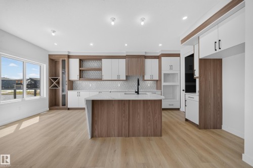 Open-concept kitchen with wood-finish flooring, a central island with a waterfall countertop, white and wood-tone cabinetry, and a herringbone tile backsplash - 8915 Mayday Way Sw, Edmonton, AB - Indoor Photo Showing Kitchen With Upgraded Kitchen