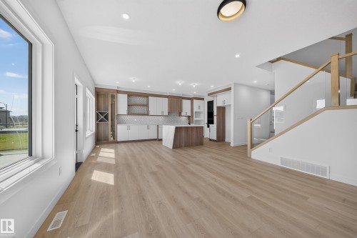 Open-concept living space with wood-finish flooring, two-tone kitchen cabinetry, kitchen island with seating, recessed lighting, and a modern glass-paneled staircase - 8915 Mayday Way Sw, Edmonton, AB - Indoor Photo Showing Other Room