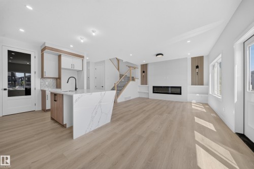 Open-concept living area with wood-finish flooring, a contemporary fireplace, and a kitchen island with a waterfall countertop - 8915 Mayday Way Sw, Edmonton, AB - Indoor Photo Showing Kitchen