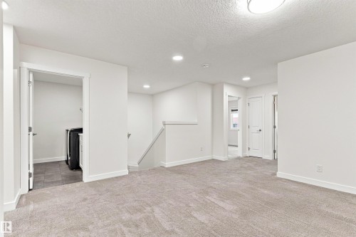 Spacious carpeted area with recessed lighting, an open doorway revealing a laundry appliance on tiled flooring, and a staircase with a white railing - 22019 82 Avenue Nw, Edmonton, AB - Indoor Photo Showing Other Room