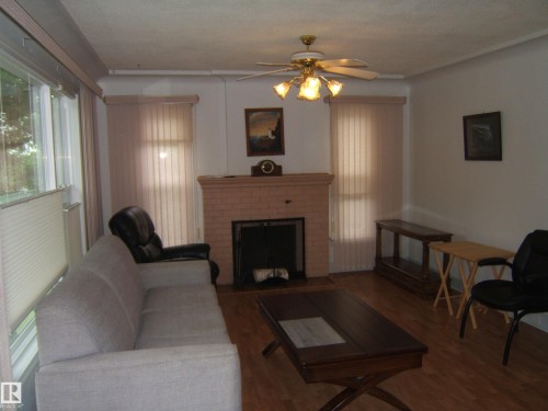 Spacious living room featuring hardwood flooring, a wood-burning fireplace with a finished mantel, ceiling fan with integrated lighting, and vertical blinds at multiple windows allowing natural light throughout the room - 4809 47A Street, Two Hills, AB - Indoor Photo Showing Living Room With Fireplace