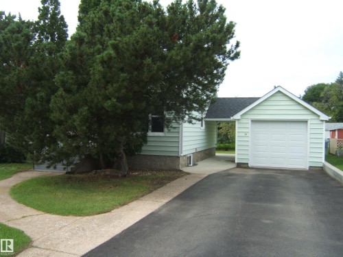 Light green horizontal siding with white trim, attached single-car garage, paved driveway, concrete walkway, and mature evergreen landscaping - 4809 47A Street, Two Hills, AB - Outdoor
