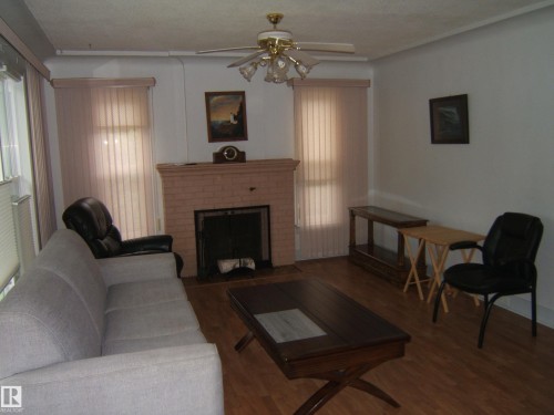 Living area featuring a brick fireplace, wood-finish flooring, ceiling fan with integrated lighting, and vertical window blinds - 4809 47A Street, Two Hills, AB - Indoor Photo Showing Living Room With Fireplace