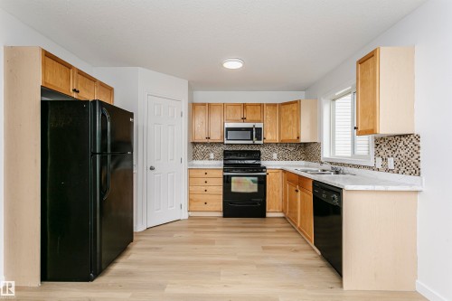 Kitchen featuring natural wood cabinetry, light-toned wood-finish flooring, a mosaic tile backsplash, white countertops, and stainless steel appliances - 37 Suntree Promenade, Leduc, AB - Indoor Photo Showing Kitchen With Double Sink