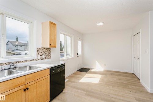Kitchen featuring wood-finish cabinetry, a double basin stainless steel sink, black dishwasher, mosaic tile backsplash, and light-tone wood-finish flooring - 37 Suntree Promenade, Leduc, AB - Indoor Photo Showing Kitchen With Double Sink