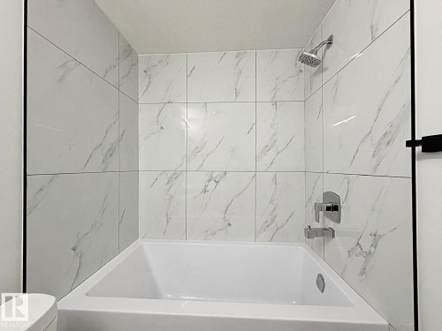 Bathtub with a shower system featuring a handheld showerhead, chrome fixtures, and white tile surround with subtle gray veining - 1312 35 Street Nw, Edmonton, AB 