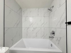 Bathtub with a shower system featuring a handheld showerhead, chrome fixtures, and white tile surround with subtle gray veining - 