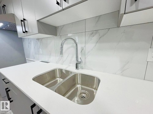 Modern kitchen featuring a double-basin stainless steel sink, pull-down faucet, white countertop, white and gray patterned tile backsplash, and light-toned cabinetry with black hardware - 1312 35 Street Nw, Edmonton, AB 
