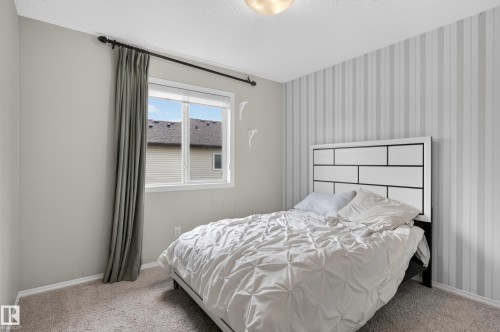 Serene primary bedroom featuring vertical striped wallpaper, a metal-frame bed with geometric headboard detailing, carpeted flooring, and abundant natural light from a dual-pane window with coordinating gray drapery - 17 Spruce Ridge Cove, Spruce Grove, AB - Indoor Photo Showing Bedroom
