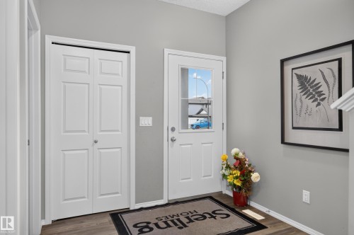 Welcoming entryway featuring dual white six-panel closet doors, a white entry door with glass panel and transom window, wood-finish flooring, soft gray walls, and framed botanical artwork - 17 Spruce Ridge Cove, Spruce Grove, AB - Indoor Photo Showing Other Room