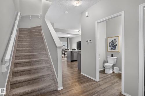 Open-concept main floor featuring wood-finish flooring throughout, a staircase with white railings and gray-toned treads, and a half bath with white cabinetry and framed artwork - 17 Spruce Ridge Cove, Spruce Grove, AB - Indoor Photo Showing Other Room