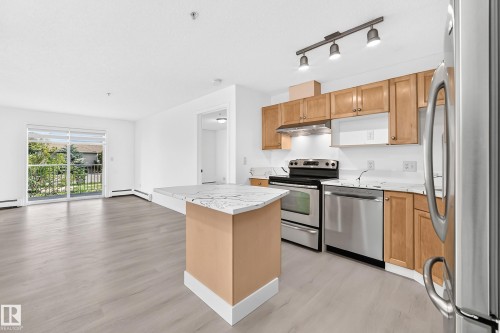 Open-concept living area featuring light wood-finish flooring, white walls, and a kitchen island with a waterfall countertop - #214 245 Edwards Dr Sw, Edmonton, AB - Indoor Photo Showing Kitchen With Double Sink